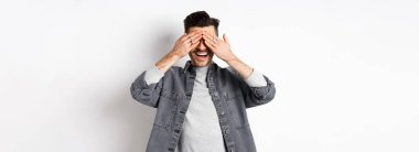 Yakışıklı mutlu adam gözlerini kapar ve şaşırmayı bekler, heyecanla gülümser, beyaz arka planda sıradan kıyafetler giyer..