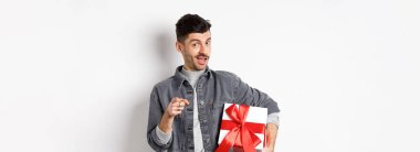 Sevgililer günü için hediye alan romantik adamın portresi ve seni işaret eden, özel alışverişe davet eden, beyaz arka planda hediyeyle duran.