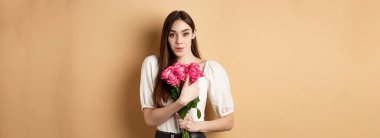 Sevgililer Günü. Şaşırmış hassas kız çiçekler için teşekkür ediyor ve gülümsüyor, kırmızı güller tutuyor ve gülümsüyor minnettar, bej arka planda duruyor..