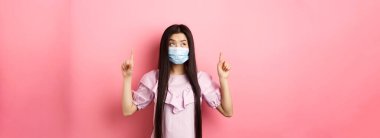Coronavirus, karantina ve yaşam tarzı konsepti. Tıbbi maskeli sevimli Koreli kız parmaklarını kaldırmış, en iyi reklamlara bakıyor, pembe arka planda duruyor..