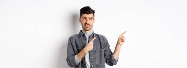 Şüpheci ve şüpheci genç adam sol logoyu işaret ediyor, nefret ve şaşkınlıkla, beyaz arka planda duruyor..