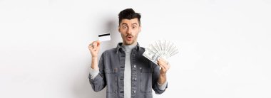 Elinde plastik kredi kartı ve dolar banknotları olan heyecanlı adam beyaz arka planda eğleniyor..