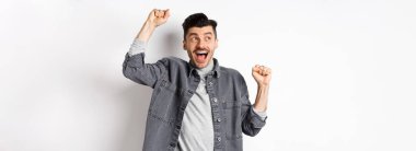 Heyecanlı genç adam tezahürat yapıyor ve yana bakıyor, ödül piyangosunu kazanıyor, iyi bir anlaşma yapıyor ve kutluyor, amacına ulaşıyor veya başarıyor, zafer dansı yapıyor ve evet diye bağırıyor, beyaz arka plan.