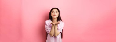 Sevimli ve romantik Asyalı kız kameraya hava öpücüğü veriyor, gözlerini kapatıyor ve nazikçe gülümsüyor, pembe arka plana karşı elbise içinde duruyor..