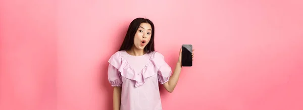 Online shopping. Excited asian woman say wow, showing empty smartphone screen and looking amazed at camera, standing against pink background.