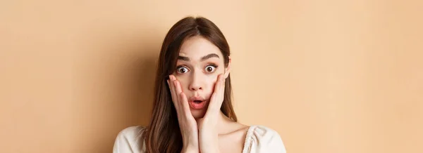 Face of surprised girl saying wow, touching cheeks and staring excited at camera, standing on beige background.