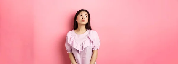 Dreamy and sad asian woman look at upper left corner logo, standing pensive and gloomy in pink romantic dress, white background.
