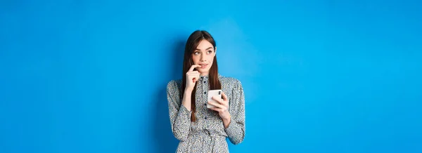 Pensive cute woman thinking how to answer on message, looking aside thoughtful and holding smartphone, standing in dress on blue background.