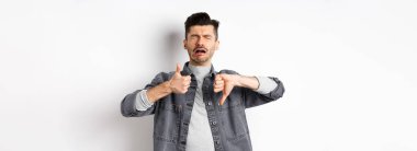 Üzgün adam ağlıyor ve baş parmağını kaldırıyor, mızmızlanıyor ve şikayet ediyor, beyaz arka planda hayal kırıklığına uğruyor..