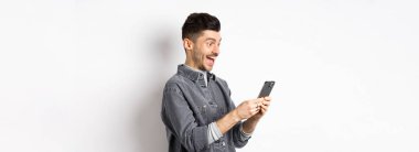 Telefonda mesaj okuyan, akıllı telefon ekranında mutlu görünen, beyaz arka planda duran heyecanlı yakışıklı adamın profil portresi.