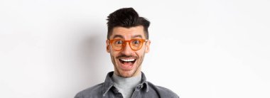 Moda gözlüklü heyecanlı yakışıklı adam harika bir tanıtıma bakıyor, gülümsüyor, beyaz arka planda mutlu duruyor..