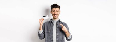 Plastik kredi kartını işaret eden bıyıklı, heyecanlı yakışıklı adam, gülümsüyor, iyi bir anlaşma öneriyor, beyaz arka planda duruyor..