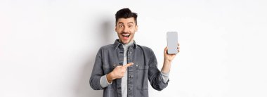 Heyecanlı yakışıklı adam boş akıllı telefon ekranı gösteriyor ve gülümsüyor, şuraya bakın, jest, beyaz arka planda duruyor..