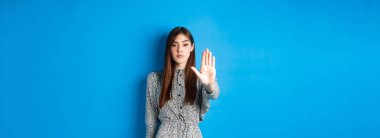 Ciddi ve kendine güvenen, uzun saçlı ve elbiseli bir kadın, elini uzat ve hayır de, jesti durdur, kötü hareketi yasakla, mavi arka planda dur de..