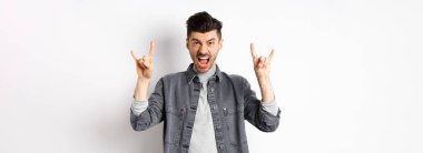 Salla gitsin. Küstah genç adam bağırıyor ve heavy metal boynuz işareti gösteriyor, harika bir konsere katılıyor, eğleniyor, beyaz arka planda duruyor..