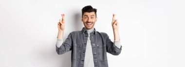 İyimser genç erkek, hayallerinin gerçekleşmesini umuyor, parmaklarını çapraz tutuyor, gülümsüyor ve iyi haberler bekliyor, beyaz arka planda duruyor..