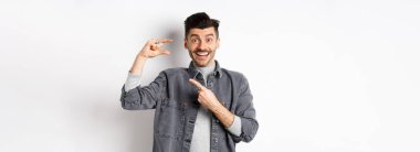 Heyecanlı adam küçük boy gösteriyor, küçük bir şeyi işaret ediyor ve gülümsüyor, beyaz arka planda duruyor..