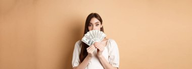 Dolarlar gösteren ve kaşlarını kaldıran heyecanlı kız hayret içinde, para kazanıyor, bej arka planda duruyor..