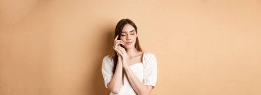 Şefkatli ve romantik bir kadın gözlerini kapatır, yumuşak yumuşak tenine dokunur, bej arka planda dururken rüya gibi gülümser..