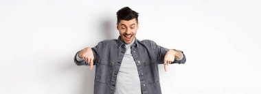 Heyecanlı Avrupalı adam aşağı bakıyor ve işaret ediyor, reklamlarda mutlu görünüyor, beyaz arka planda kot ceketle duruyor..