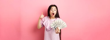 Heyecanlı genç kız elinde büyük miktarda para tutuyor, dolarları ve başparmakları gösteriyor, pembe arka planda şaşkın şaşkın duruyor..