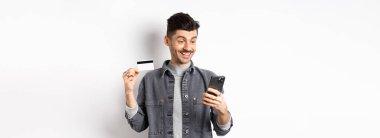 İnternetten alışveriş konsepti. Gülümseyen adam cep telefonuyla ödeme yapıyor, plastik kredi kartı gösteriyor, internetten alıyor, beyaz arka plan.