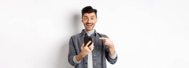 Heyecanlı genç adam telefonda iyi haberler alıyor, akıllı telefona işaret ediyor ve mutlu bir şekilde gülümsüyor, beyaz arka planda duruyor..