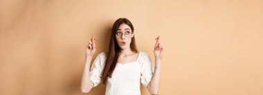 Heyecanlı kız umut rüyası gerçekleşiyor, parmaklarını çapraz yapıp dua ediyor, bej arka planda duruyor..