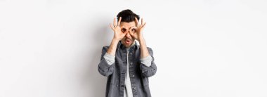 Komik adam el dürbününden bakıyor, muhteşem tanıtımlara bakıyor, hayret ediyor ve kameraya bakıyor, heyecanlı, beyaz arka planda duruyor..