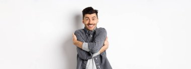 Mutlu yakışıklı adam kendini kucaklıyor ve gülümsüyor, kendi vücudunu kucaklıyor, gri ceketle beyaz arka planda duruyor..