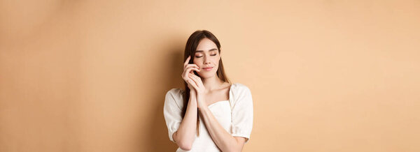 Tender and romantic woman close eyes, gently touching soft smooth skin, smiling dreamy while standing against beige background.