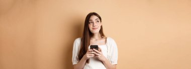 Telefonla düşünen romantik bir kız, kenara bakıyor ve gülümsüyor, akıllı telefonda flört uygulaması kullanıyor, bej arka planda duruyor..