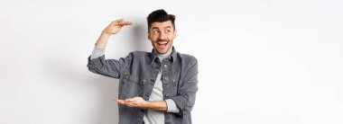 Heyecanlı adam logoya bakıyor ve büyük bir şey gösteriyor, büyük bir nesne şekillendiriyor ve şaşkın bir şekilde gülümsüyor, beyaz arka planda kot ceketle duruyor..