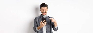 Heyecanlı yakışıklı adam telefonda güzel haberler veriyor, akıllı telefona işaret ediyor ve mutlu gülümsüyor, beyaz arka plana karşı duruyor..