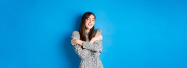 Gülümseyen romantik kız kendine sarılıyor, gözlerini kapıyor ve mutlu oluyor, mavi arka planda duruyor..