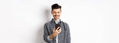 Yakışıklı adam telefonda mesaj okuyor ve gülümsüyor. Akıllı telefon tutan ve beyaz arka planda duran, ekrana bakan adam..