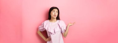 Cahil Asyalı kadın yüzünü çevirmiş, omuz silkiyor, elini kaldırıyor. Ne jesti, pembe arka planda rahatsız edilmeden duruyor..