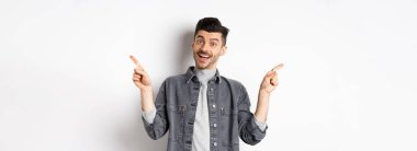 Gülümseyen neşeli adam parmaklarını yana doğru gösteriyor, sağa sola bakıyor, kameraya mutlu bakıyor, beyaz arka planda duruyor..