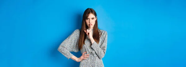 Angry and bossy young woman tell to keep quiet, show shush taboo sign and frowning upset with bad behaviour, standing on blue background.