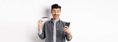 İnternetten alışveriş konsepti. İnternetten alışveriş yapan heyecanlı bir adam, plastik kredi kartı ve akıllı telefonla kolay satın alma, kameraya mutlu bir gülümseme, beyaz arka plan..