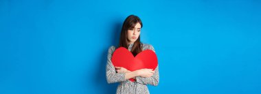 Sevgililer Günü. Yalnız bekar bir kız büyük kırmızı kalbi kucaklıyor ve üzgün üzgün bakıyor, mavi arka planda kasvetli duruyor..