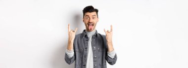 Heyecanlı komik adam dil gösteriyor ve ağır metal boynuzları işareti yapıyor, eğleniyor, eğleniyor, beyaz arka planda duruyor..