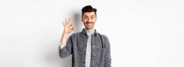 Gülümseyen yakışıklı adam iyi ürün gibi tamam işareti gösteriyor, reklam öneriyor, beyaz arka planda tatmin olmuş duruyor..