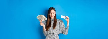 Alışverişe. Elbiseli güzel kız yana bakıyor ve gülümsüyor dalgın dalgın, para ve kredi kartı gösteriyor, mavi arka planda duruyor..