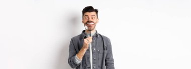 Yakışıklı, pozitif bir adam büyüteçle beyaz, mükemmel bir gülümseme ve şaşı bakışlar sergiliyor, komik suratlar yapıyor, beyaz arka plan..