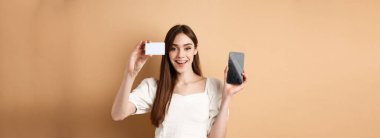 Plastik kredi kartı ve boş cep telefonu ekranı gösteren neşeli genç bir kadın. Kameraya gülümsüyor, bej arka planda duruyor..