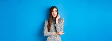 Uzun doğal saçlı, eski moda elbiseler giyen, düşünceli yüzüyle sol tarafa bakan, mavi arka planda duran düşünceli beyaz kız..