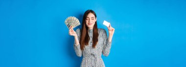 Alışverişe. Mutlu gülümseyen kadın, gözleri kapalı, para ve kredi kartı gösteren, mavi arka planda hayaller kuran mutlu bir kadın..