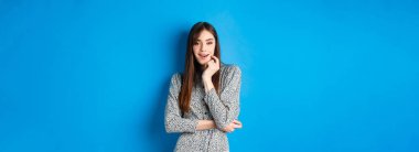 Uzun elbiseli, çekici kız kameraya bakıyor, mavi arka planda duruyor..