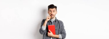 Burnunu karıştıran ve kitap tutan genç adam, kameraya aptal gibi bak, beyaz arka planda duruyor..